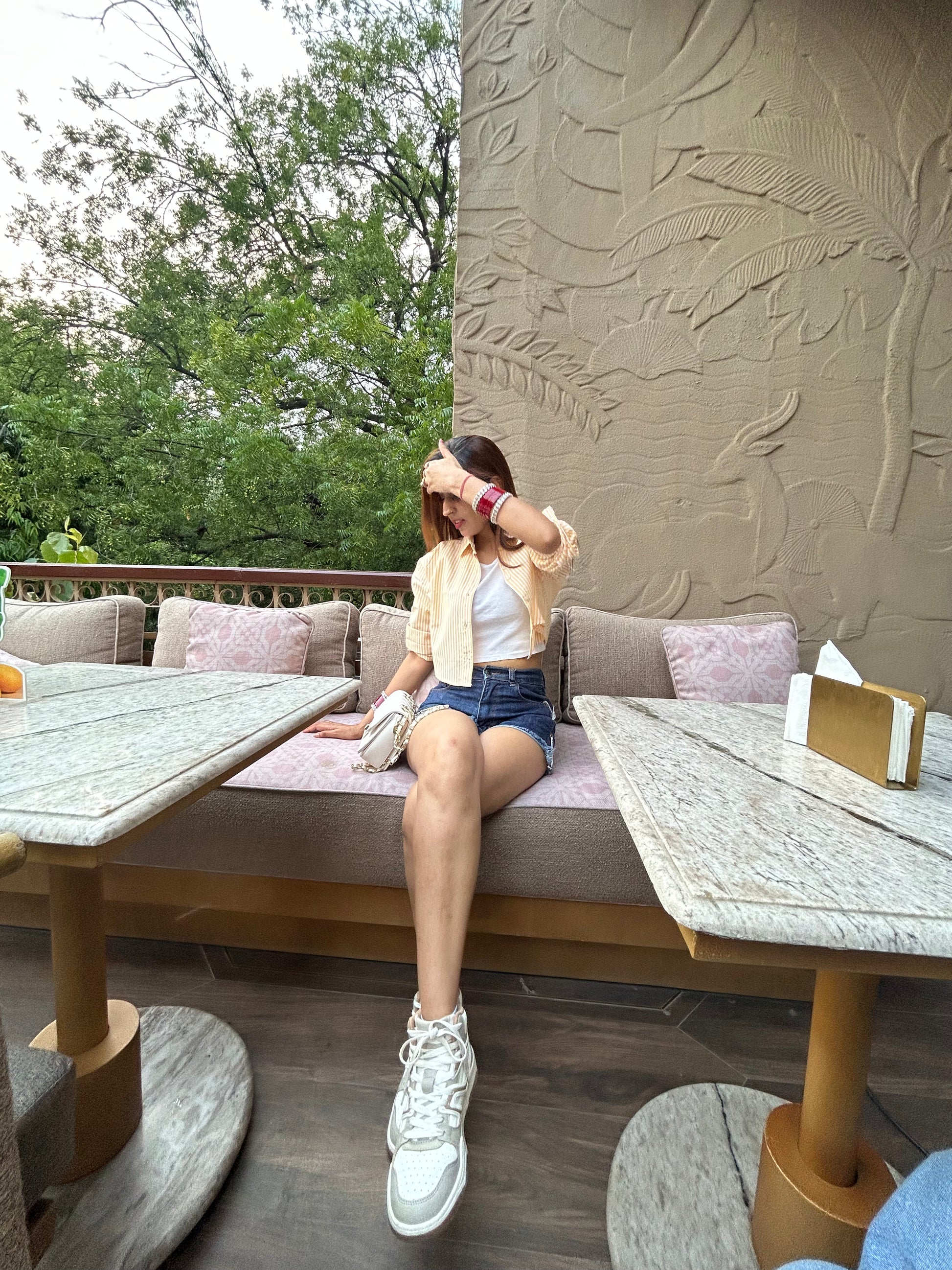 Woman sitting on a bench in an outdoor setting wearing a yellow and white striped crop shirt with a white crop top underneath it, with trees and a textured wall.