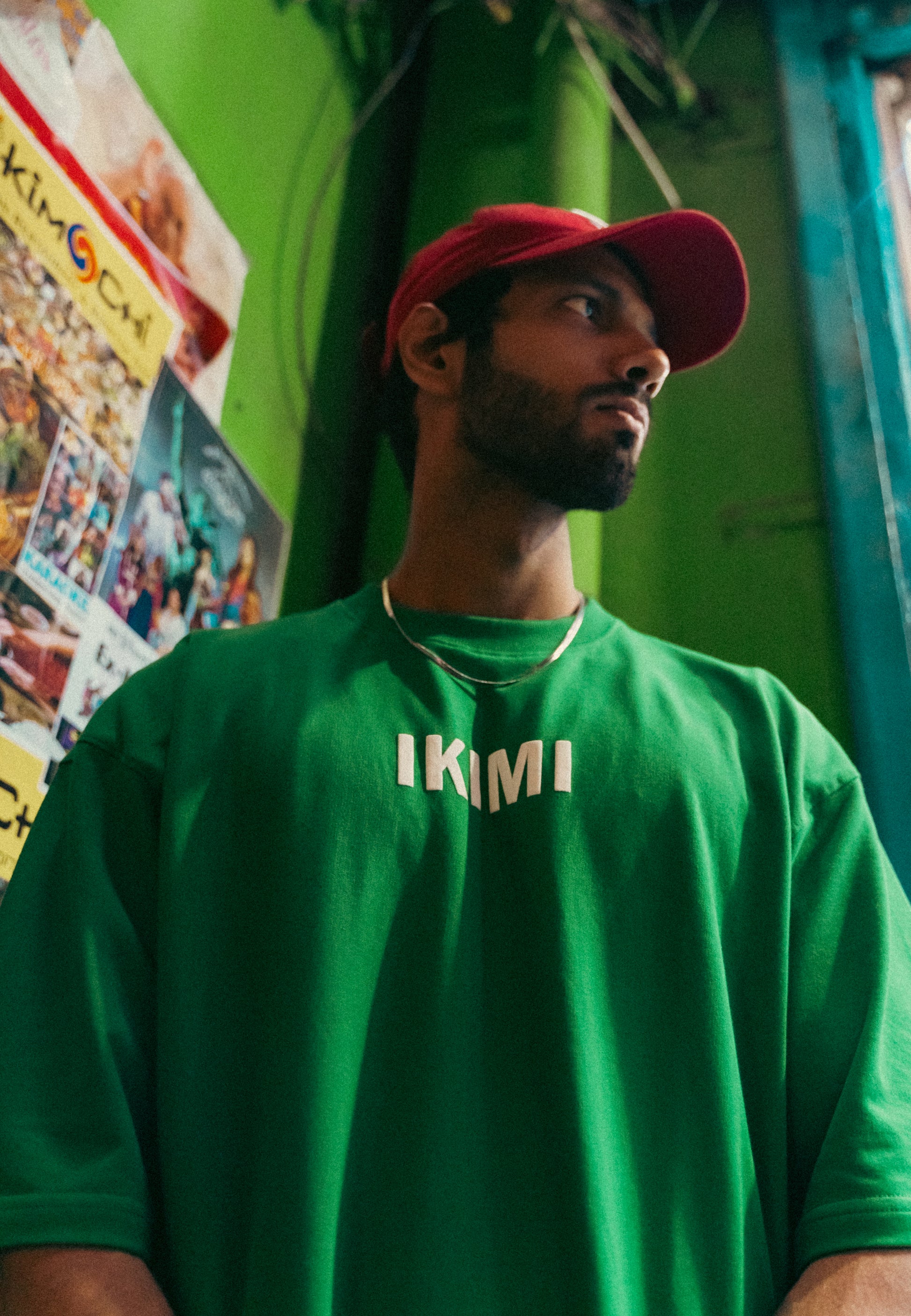 A person wearing a green round neck T-shirt with the brand name 'IKIMI' on it, paired with a red cap.