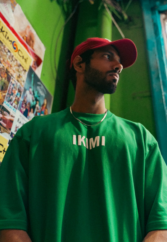A person wearing a green round neck T-shirt with the brand name 'IKIMI' on it, paired with a red cap.