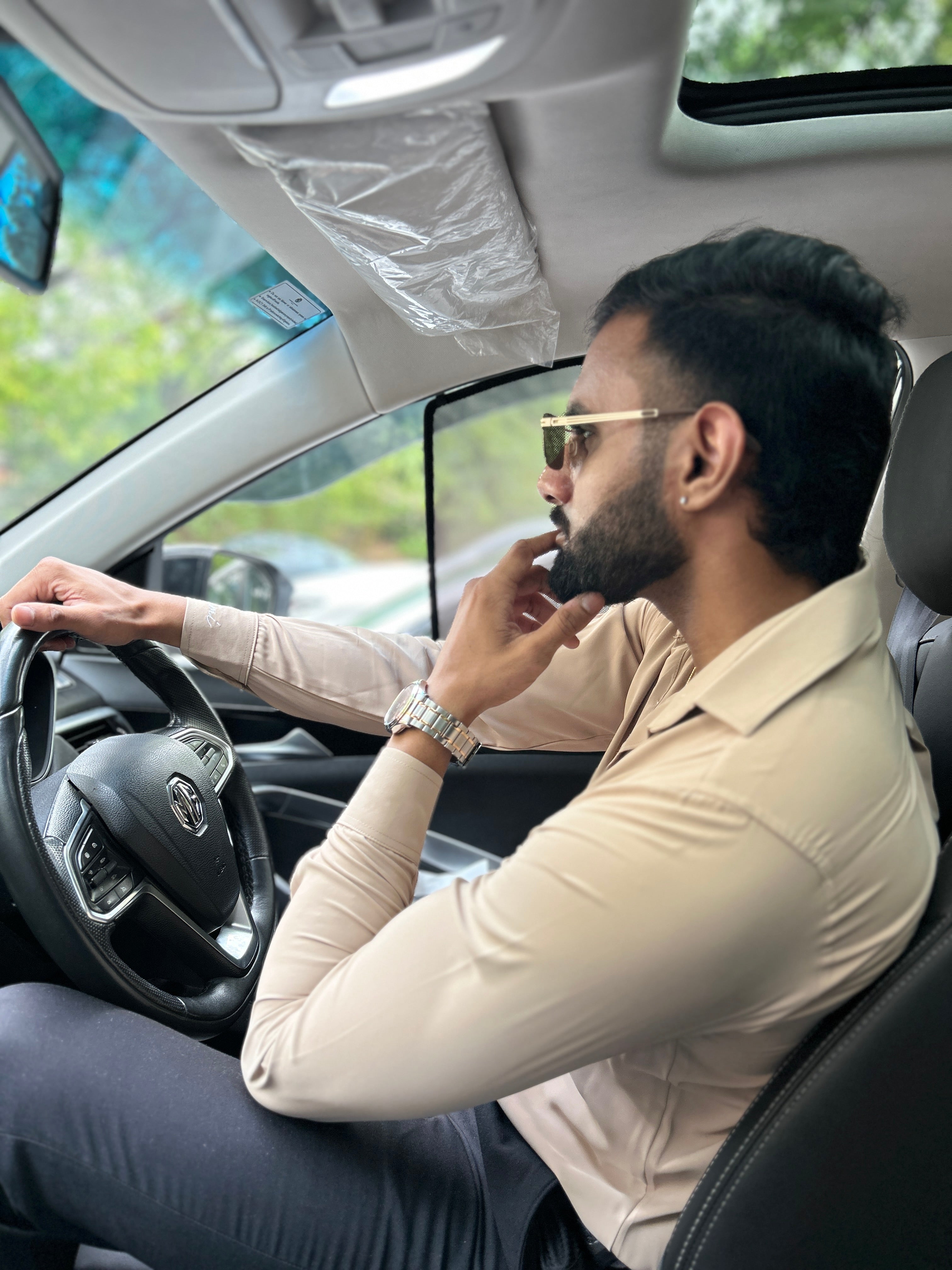 Man driving a car with sunglasses on, wearing a beige shirt and watch.